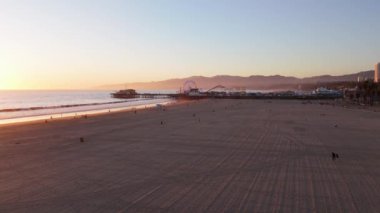 Hava 4k. Alacakaranlık okyanusu dalgaları, klasik dönme dolap, Santa Monica Pasifik Okyanusu sahilindeki iskeledeki eğlence parkı, altın gün batımında. Kaliforniya 'nın alacakaranlıkta parlayan simgesi, Los Angeles