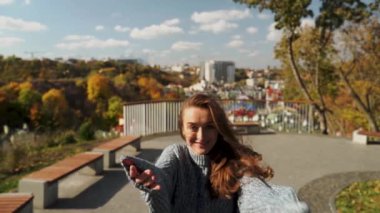 Genç bir kadın, canlı renkler ve güzel bir şehir zeminiyle çevrili pitoresk bir sonbahar parkında neşeyle kutlama yapıyor. Sıcak bir kazağa sarılmış neşeli bir kadın manzaranın tadını çıkarıyor.