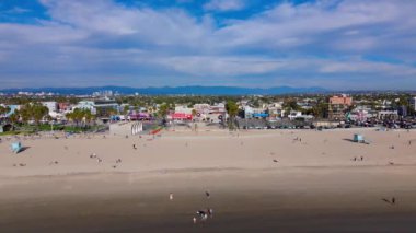 Canlı bir plaj sahnesi ziyaretçilerin sıcak güneşin ve bulutlu bir gökyüzünün altındaki yumuşak kumların keyfini çıkardığını gösteriyor. Venice plajı, Los Angeles, ABD. 2024. Yüksek kalite 4k görüntü