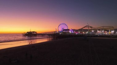 Santa Monica, LOS ANGELES, CALIFORNIA, ABD, Ekim 10, 2024. Göz kamaştırıcı bir dönme dolap ve sıcak renklerle aydınlatılmış heyecan verici lunapark treniyle hareketli bir iskelede gün batımını deneyin. Yüksek