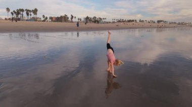 Su ve palmiye ağaçlarında yansımaları olan güzel kumlu bir plajda amuda kalkan bir çocuk. LOS ANGELES, CALIFORNIA, ABD, 10 Ekim 2024. Ağır çekim. Yüksek kaliteli görüntüler