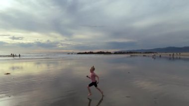 Neşeli bir çocuk, engin ve bulutlu bir gökyüzünün altındaki güzel yansıtıcı kıyı şeridi boyunca enerjik bir şekilde koşuyor. LOS ANGELES, CALIFORNIA, ABD, 10 Ekim 2024. Ağır çekim. Yüksek kaliteli görüntüler