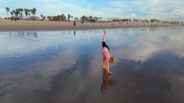 Neşeli bir kız sahilde oynuyor, güneşle amuda kalkıyor ve arkasında dalgalar sallıyor, okyanusları kucaklıyor ferahlatıcı suları kucaklıyor. LOS ANGELES, CALIFORNIA, ABD, 10 Ekim 2024. Ağır çekim. Yüksek kalite
