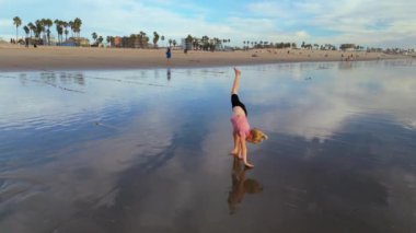 Neşeli bir kız sahilde oynuyor, güneşle amuda kalkıyor ve arkasında dalgalar sallıyor, okyanusları kucaklıyor ferahlatıcı suları kucaklıyor. LOS ANGELES, CALIFORNIA, ABD, 10 Ekim 2024. Ağır çekim. Yüksek kalite