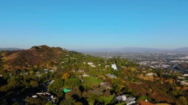 Los Angeles, Kaliforniya 'daki Hollywood Tepeleri üzerinde ilerleyen hava aracı görüntüleri. Manzaralar yemyeşil tepeler, lüks evler ve Los Angeles 'ın engin manzarasıyla berrak mavi bir gökyüzü gösteriyor.