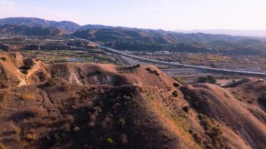 Şubat 2025 'te çekilen hava görüntüleri Kaliforniya, ABD' de kayan tepeleri ve otobanı gösteriyor. Kuru bitki örtüsüyle kaplı engebeli arazi, arka plandaki gelişmiş altyapıyla çelişiyor.
