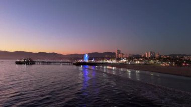 Santa Monica, LOS ANGELES, CALIFORNIA, ABD, Ekim 10, 2024. Göz kamaştırıcı bir dönme dolap ve sıcak renklerle aydınlatılmış heyecan verici lunapark treniyle hareketli bir iskelede gün batımını deneyin. Yüksek