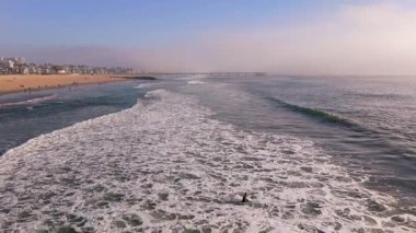 Venice Beach, Los Angeles 'ta sisli bir Kasım sabahı köpüklü okyanus dalgalarında kürek çeken bir sörfçünün hava yavaş çekim görüntüleri. Uçsuz bucaksız sahil şeridi, yumuşak dalgalar ve yumuşak güneş ışığı...