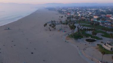 10 Ekim 2024 'te Los Angeles, Kaliforniya' daki Venice Beach 'in hava görüntüleri. Kamera geriye doğru hareket ediyor, geniş kumlu kıyı şeridini, palmiye ağaçlarını, kaykay parkını ve hareketli sahil alanını gösteriyor.