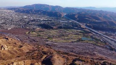 Çarpıcı hava görüntüleri, engebeli bir dağ manzarasına kusursuz bir şekilde uyum sağlayan bir şehri gösteriyor. Parlak yeşil alanları olan bir golf sahası kurak tepelerle tezat oluştururken, büyük bir karayolu kesimi