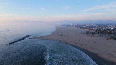 Los Angeles, Kaliforniya 'daki Venice Beach' in hava görüntüleri. İnsansız hava aracı, engin kumlu kıyı şeridini, okyanus dalgalarını ve şehir manzarasının uzak bir görüntüsünü ortaya çıkararak ilerliyor. İkonik palmiye ağaçları
