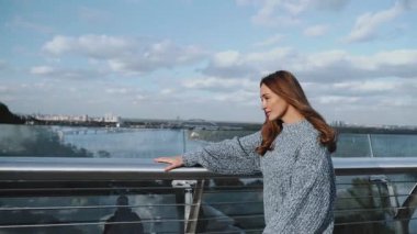 Genç bir kadın, bulutlu bir gökyüzünün altındaki dingin göl manzarasına dikkatle bakan bir korkuluğun yanında zarif bir şekilde duruyor. Şehir arka planında uzun saçlı, kalın kazaklı bir kız. Ukrayna, 2021. Yavaş