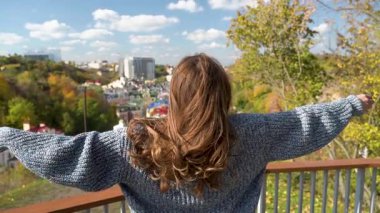 Uzun kıvırcık saçlı bir kadın bir balkonda duruyor, kazak giymiş, yüzünü kilitlerine gizliyor, hareketli şehirde özgürlüğü yansıtıyor. Kyiv, Ukrayna, 2021. Ağır çekim. Yüksek kaliteli HD