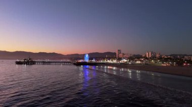 Santa Monica, LOS ANGELES, CALIFORNIA, ABD, Ekim 10, 2024. Göz kamaştırıcı bir dönme dolap ve sıcak renklerle aydınlatılmış heyecan verici lunapark treniyle hareketli bir iskelede gün batımını deneyin. Yüksek