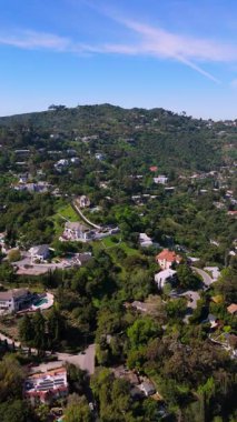 Los Angeles 'taki yeşil tepelerle çevrili lüks evleri gösteren dikey hava görüntüleri. Kamera ilerler, konut mimarisini, bereketli manzaraları ve dolambaçlı yolları ön plana çıkarır.