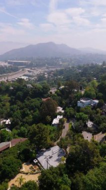 Hollywood Tepeleri 'nin üzerinden geriye doğru uçan dikey insansız hava aracı görüntüleri yemyeşil alanları, özel yamaçlardaki evleri ve uzak otoyol kavşaklarını yakalıyor. Hollywood tabelası dağın tepesinde görünür.