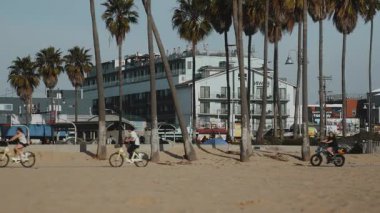 Los Angeles 'taki Venice Beach Boardwalk' ta canlı bir sahne. Palmiye ağaçları, modern binalar ve kumlu yolda bisiklet süren bisikletçilerin yer aldığı. Kentsel sahil atmosferi net bir şekilde görüntülenmiştir.