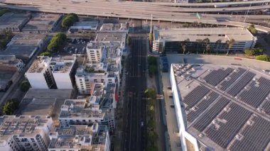 Los Angeles, Kaliforniya şehir merkezinin dinamik tepeden aşağı manzarası, şehir sokakları, modern yerleşim binaları, güneş panelli çatılar ve gün doğumunda çok katlı bir otoyol. Arabalar sürüyor.