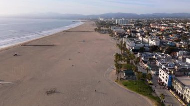 Los Angeles, Kaliforniya 'daki Venice Beach' in insansız hava aracı görüntüleri. Kamera, geniş kumlu bir sahili, Pasifik Okyanusu 'nu ve palmiye ağaçları ve modern gökdelenlerle dolu canlı bir tahta kaldırımı gözler önüne sererek ileri doğru hareket ediyor.
