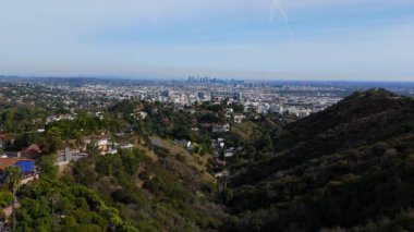Manzaralı bir kanyondan çekilen Los Angeles gökyüzü manzarası. İnsansız hava aracı ileri uçuyor, konut tepelerini, yemyeşil alanları ve parlak mavi gökyüzünün altındaki panoramik şehirleri gösteriyor.