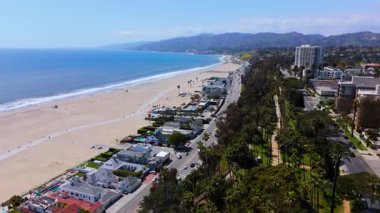 Santa Monica kıyı şeridi boyunca ilerleyen hava aracı görüntüleri, sahili, okyanus dalgalarını, palmiyeli caddeleri, kıyı otoyolunu ve lüks gayrimenkulleri yakalıyor. Manzarada yayalar da var.