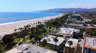 Yavaş çekim drone görüntüleri Santa Monica, California 'da sahil yolunda ilerlerken Pasifik Okyanusu' nun, palmiyeli parkın ve sahil malikanelerinin pitoresk manzarasını gözler önüne seriyor. Sahne