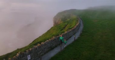 Havadan. Turistler Moher 'in uçurumundaki puslu yolu takip ediyorlar. Anne ve çocuk, Moher Kayalıkları 'ndaki uçurumlar boyunca turist yolunda bir uçurum boyunca yürüyorlar. Güzel gün batımı. Country Clare.