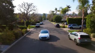 Hava aracı görüntüleri, Studio City, Los Angeles, California, ABD 'deki manzaralı yerleşim yerlerini gösteriyor. Beyaz bir araba sakin bir banliyö caddesi boyunca palmiye ağaçları ve yemyeşil ağaçlarla kaplı.