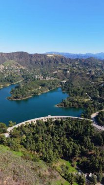 Hollywood Tabelasının muhteşem insansız hava aracı görüntüsü. Tepelerin üstünde Hollywood Gölü ve Mulholland Barajı ön planda. Berrak mavi gökyüzünün altında yeşilliklerle çevrili ikonik Los Angeles simgelerini yakalar