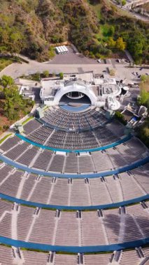 Kaliforniya tepelerinde yuva yapmış büyük bir açık hava amfitiyatrosunun dikey drone görüntüsü simetrik mimari, katmanlı oturma ve doğal çevreyi gösteriyor. Kamera tepeden tırnağa görüntüler.