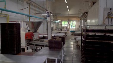 Geleneksel bir endüstriyel pastanenin içinde metal işyerleri, fırın tepsileri, hamur işleme ekipmanları ve arka planda bir işçinin yer aldığı el kamerası görüntüsü. Üretim alanı tepsilerle dolu.