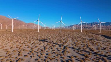 Düzinelerce dönen türbinle birlikte geniş bir çöl rüzgâr çiftliğinin üzerinden havadan sola doğru görüntüler geliyor. Beyaz kulelerin temiz çizgileri, kuru altın arazi ve dramatik dağ ile tezat oluşturuyor.