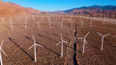 Drone, güneş doğarken çölde geniş bir rüzgar çiftliğini yakalıyor. Düzinelerce modern rüzgar türbini ufukta bitip tükenmeden uzanıyor. Sabah ışığı kurak arazide uzun gölgeler bırakır.