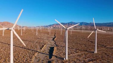 İlk ışıkta yakalanan büyük bir rüzgar enerjisi tesisinin geniş bir hava görüntüsü. Kamera yatay olarak sağdan sola süzülüyor. Sonsuz bir rüzgar türbinleri ızgarası boyunca uyum içinde dönüyor.