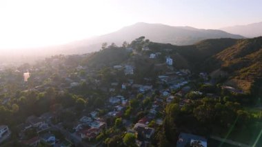 Los Angeles, Kaliforniya 'daki yerleşim yerlerinin havadan çekilmiş görüntüleri. İnsansız hava aracı, bereketli ağaçlar ve dramatik dağlarla çevrili dolambaçlı yolların ve tepe kenarındaki evlerin üzerinde uçuyor.