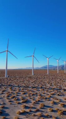 Palm Springs, Kaliforniya 'daki çöl manzarasında açık mavi gökyüzü ve arka planda uzak dağlarla birlikte rüzgar türbinlerinin dikey görüntüsü. Kamera yavaşça ileri doğru hareket ediyor.