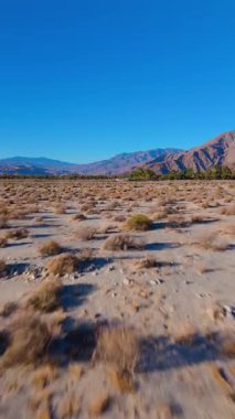 Dikey İHA görüntüleri, Palm Springs, Kaliforniya 'daki kurak çöl arazisinde, açık mavi gökyüzünün altındaki uzak sıradağlarla hızla uçuyor. Kamera yüksek hızda ileri doğru hareket ediyor.