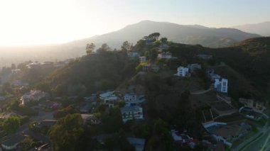 Los Angeles, Kaliforniya 'da günbatımında tepeler boyunca inşa edilen lüks konutların hava manzarası. Kamera, sıcak altın ışığın manzarayı yıkadığı sessiz muhitte ilerliyor.
