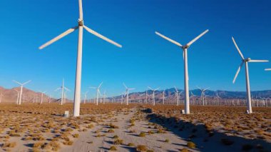 İnsansız hava aracı, büyük rüzgar türbinleri arasında alçak irtifada uçarak kuru bir çöl ovasında hareket ve ölçekleme hissi yaratır. Parlak gün ışığı ve açık gökyüzü temiz enerji kavramını vurguluyor ve