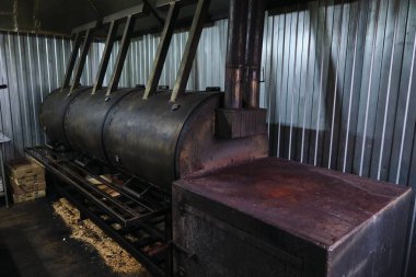 A large, industrial smoker used for preparing and cooking meat, positioned indoors against a corrugated metal wall. The smoker is black and cylindrical with multiple chambers for smoking different types of meats simultaneously. clipart