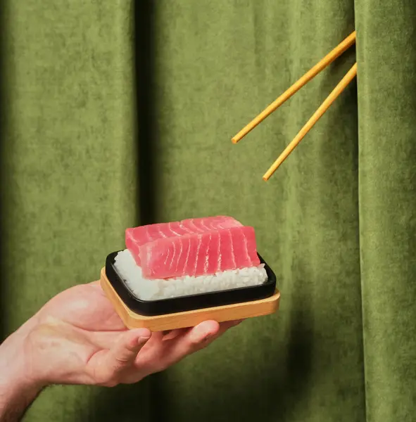 stock image A hand holds a sushi platter featuring a slice of fresh tuna placed atop sushi rice, set against a rich green background, creating an inviting culinary atmosphere.