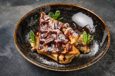 Çikolata soslu lezzetli waffle ve taze nane yapraklarıyla süslenmiş, koyu arkaplanda şık bir tabakta servis ediliyor..