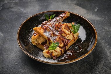 Çikolata soslu lezzetli waffle ve taze nane yapraklarıyla süslenmiş, koyu arkaplanda şık bir tabakta servis ediliyor..