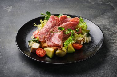 Koyu bir tabakta çeşitli et, peynir ve taze sebzelerle servis edilen gurme bir antipasto tabağının yakın plan görüntüsü. Yemek taze ve lezzetli..