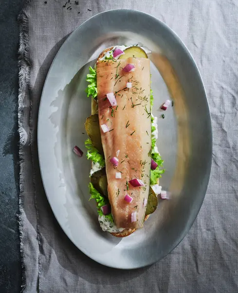 stock image A beautifully arranged herring sandwich features crisp greens, pickles, and red onion on a soft bun, all presented on an elegant silver platter, evoking gourmet dining.