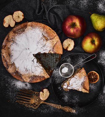 Taze pişmiş bir elma ve armut keki, toz şekerle kaplanmış koyu bir kumaşın üzerinde oturur. Dilimler kesilir ve tam meyve ve kurutulmuş elma halkalarının yanına yerleştirilir..