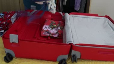 Kadın elleri kırmızı bir bavula giysiler yerleştiriyor ve düzenliyor, farklı renk ve desenlerden elbiseler giyiyor, yatakta açık, mavi ve beyaz renkler. Seyahat kavramları