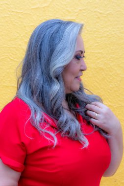 Portrait of a woman with her head turned down and her long gray hair on her chest on a yellow background, in a red dress with a black belt, modern make-up in warm colors