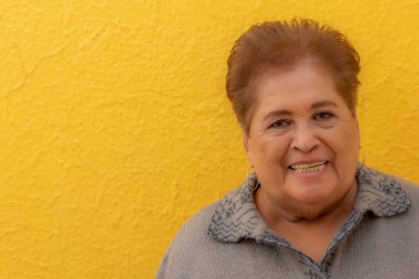 Portrait of smiling elderly woman with wide smile against yellow background, modern professional make-up, warm tones, short hair with brown tint, gray blouse. Concept of elegance and old age fashion
