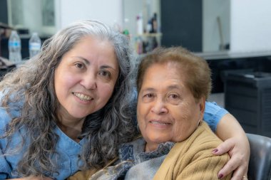 Two Latin American women mother and daughter embracing smiling and happy, looking at camera, no makeup, casual clothes, gray hair, painted with brown dye, part of a beauty salon in blurred background